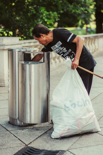 A large collection of overflowing rubbish bags, cardboard boxes, and loose paper scattered around a metal litter bin in an urban parking area. The rubbish includes black and transparent plastic bags f