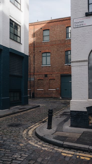 The image depicts a narrow cobbled street corner in an urban area, with a white building on the right displaying a street sign reading 'Hoxton Square' and a white-painted base with black detailing, ad