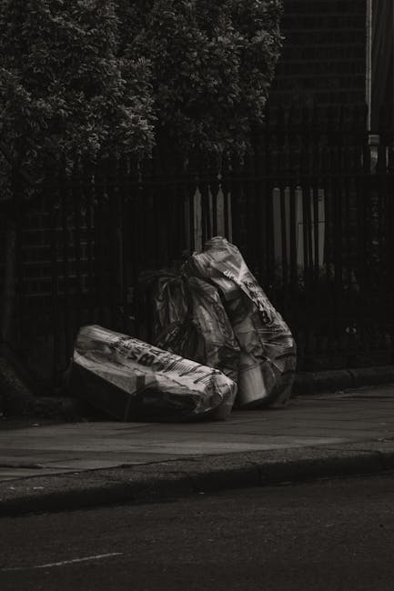 Three large black plastic garbage bags, filled and tied at the top, are leaning against a wooden fence along the side of a paved street. The bags appear to be used for rubbish collection, with some su